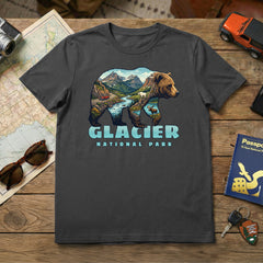 The Glacier – Grizzly Mashup T-Shirt in dark gray displays a grizzly bear graphic filled with mountains, trees, and a river, plus “Glacier National Park” below. Shown with a map, sunglasses, passport, toy car, keys, and camera.
