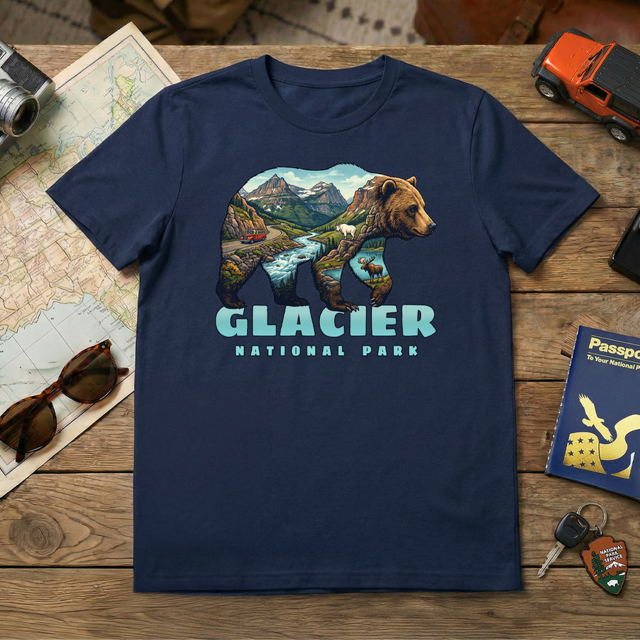 The Glacier – Grizzly Mashup T-Shirt is a navy blue tee featuring a bear graphic with mountains, trees, and a river, plus "Glacier National Park" text. The shirt is pictured with travel items like a map, passport, sunglasses, keys, and a toy car.