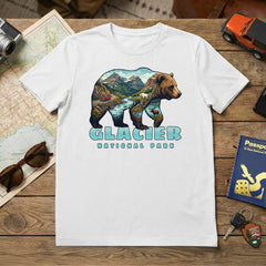A white Glacier – Grizzly Mashup T-Shirt with a bear, mountain, lake, and forest graphic sits on a wooden table beside travel essentials: map, sunglasses, camera, toy car, passport, and notebook.