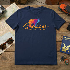A Glacier – Mountain Goat Script T-Shirt in navy blue, featuring a colorful mountain goat graphic, sits on a wooden table with travel items—a perfect Montana souvenir.