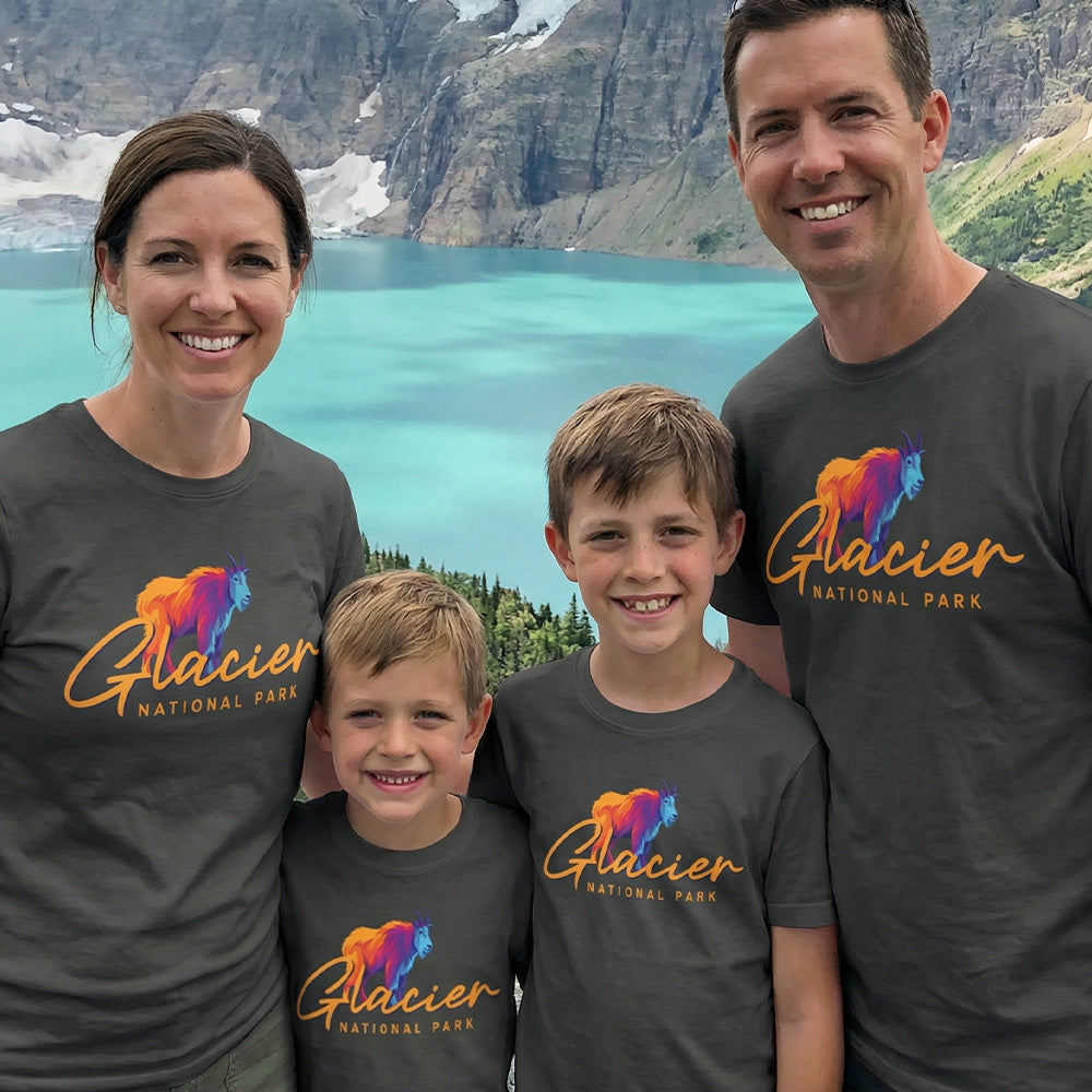 A family of four smiles by a lake and mountains, all wearing matching Glacier – Mountain Goat Script T-Shirts, which feature a colorful mountain goat graphic as a Montana souvenir.