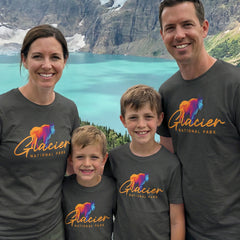 A family of four smiles by a lake and mountains, all wearing matching Glacier – Mountain Goat Script T-Shirts, which feature a colorful mountain goat graphic as a Montana souvenir.