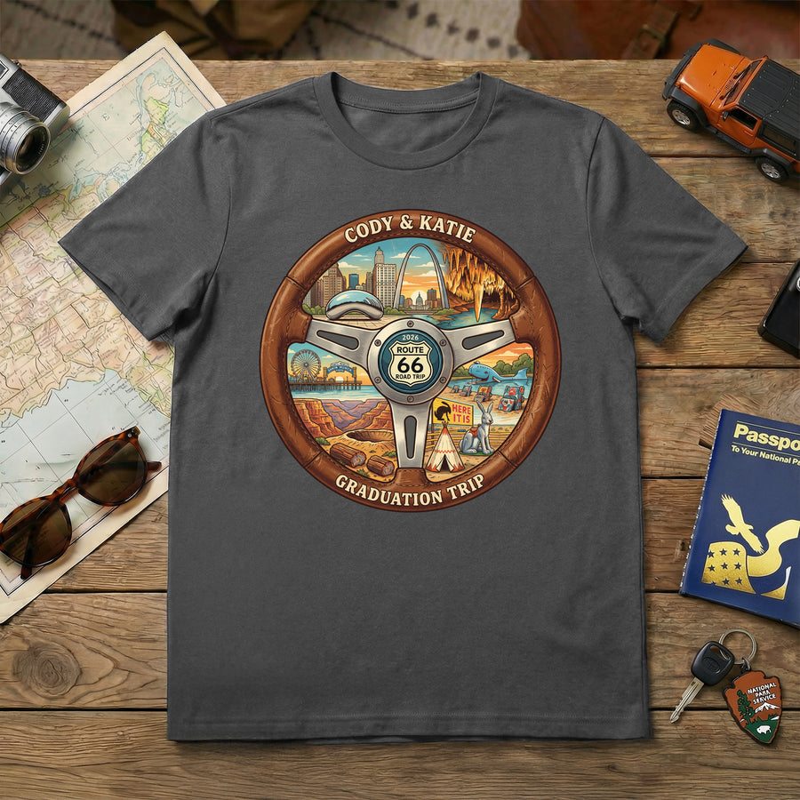 Gray t-shirt with a Route 66 steering wheel design on a wooden surface with travel-themed items.